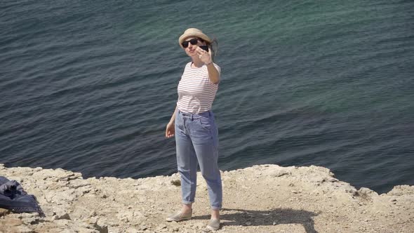 A Woman in a Straw Hat and Sunglasses Takes a Photo By the Sea alt