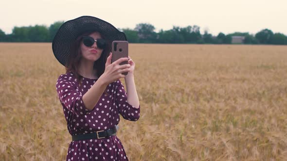Attractive Fun Hippie Woman in the Wheat Field at Sunset Making a Selfie with Smartphone alt