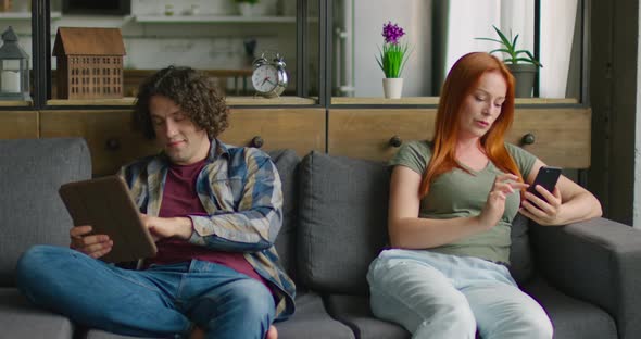 Young Man and Woman Are Sitting on Sofa, Relaxing, Using Their Gadgets alt