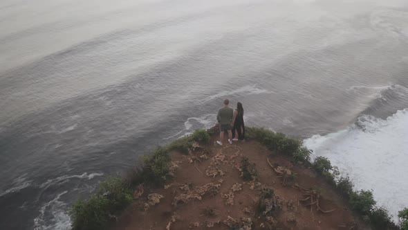 Drone View of Man and Woman Standing in Edge of Rock Looking at Sea alt