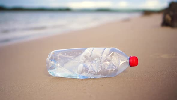 Slow motion close-up of plastic bottle being picked up from calm sandy beach. Water in background. alt