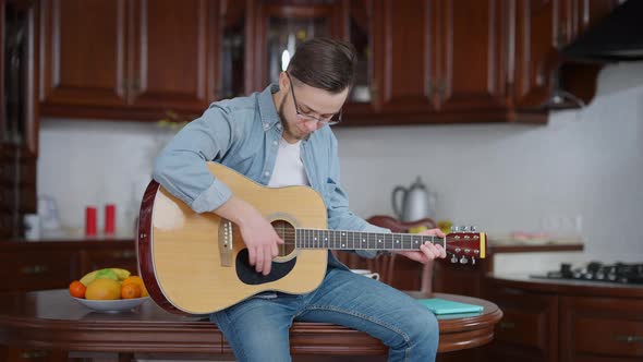 Young Romantic Caucasian Man in Eyeglasses Playing Guitar Sitting on Kitchen Table at Home alt