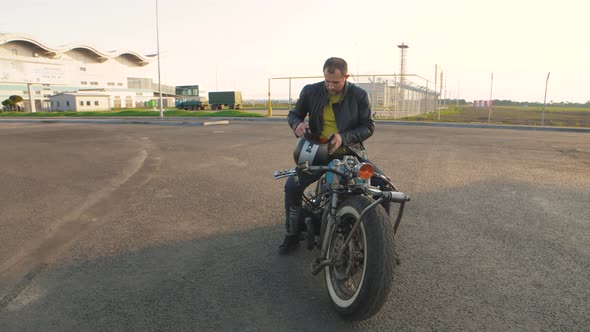 Young Man Biker with Custom Bobber Motorcycle on Street at Sunset alt