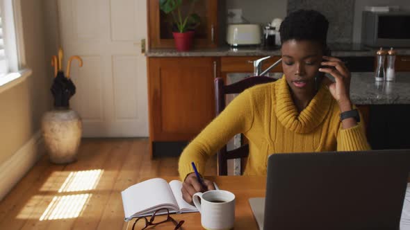 African american woman talking on smartphone and taking notes while working from home alt