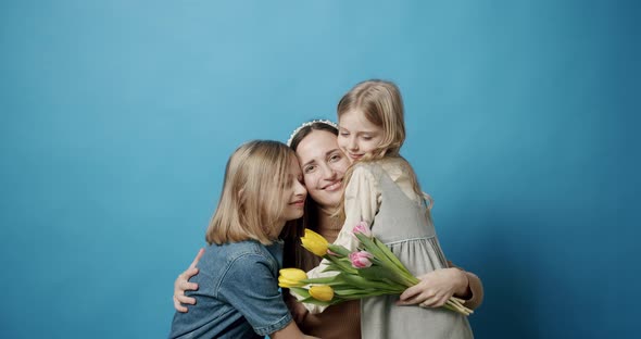 Mom Hugs Daughters and Kisses with Flowers Posing on a Blue Background alt
