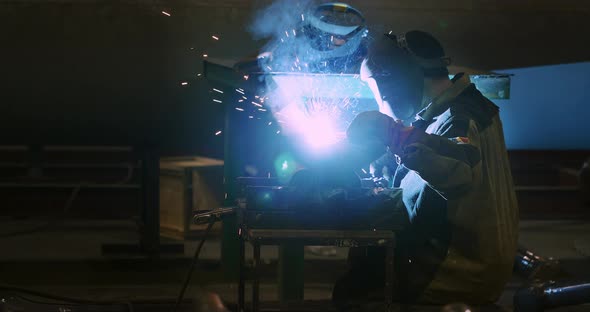 Welder in a welding helmet, in work clothes welds a pipe at the factory. Sparks alt