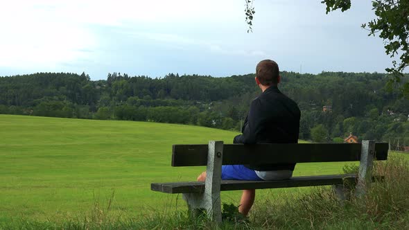 Young Man Sits on the Bench in Nature (Field with Forest) and Looks Around alt