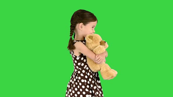 Little Girl in Polka Dot Dress Hugging Big Teddy Bear While Walking and Looking at Camera on a Green alt