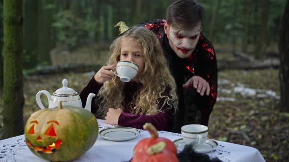 Cute Caucasian Little Girl Drinking Tea in Forest As Man with Zombie Makeup in Costume Scaring Kid alt