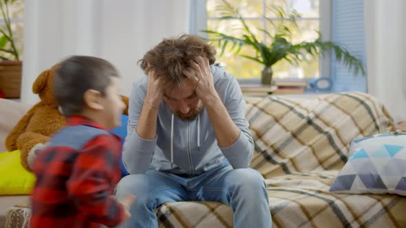 Young Man Sitting on Couch Tired of Noisy Children Running and Shouting alt