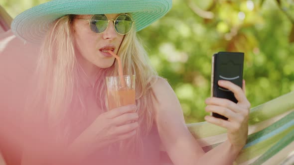 Smiling blonde woman with sunglasses using smartphone, relaxing on the hammock in garden