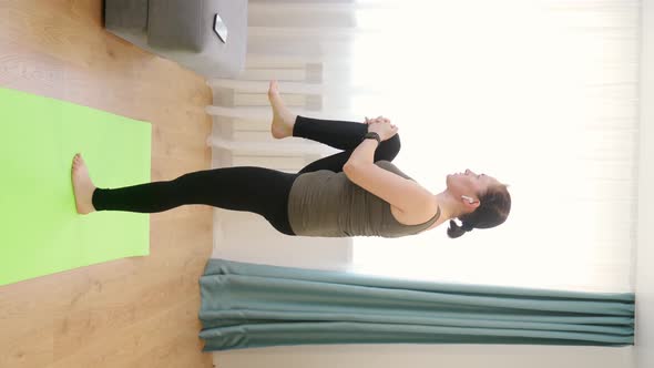 A Woman in a Tshirt Black Leggings is Doing Exercises on a Rug in a Bright Room alt