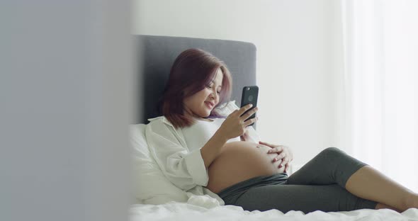 Asian pregnant wife using smartphone while lying on a bed in bedroom.