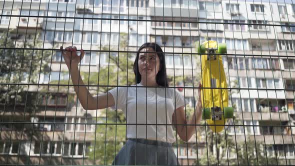 Portrait of Cute Woman with a Skateboard Looking at the Camera Standing Behind the Mesh Fence alt