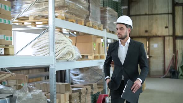 Young boss in a suit and white hard hat walks through a warehouse alt