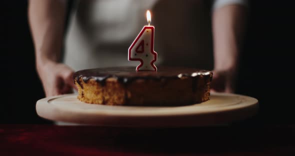 Young Woman Wearing Kitchen Apron Puts Homemade Chocolate Cake or Pie with Number 4 Burning Candle alt