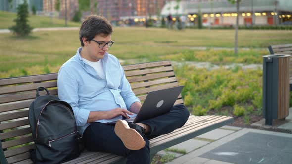 Young Man Using Laptop in Park Sitting on a Bench on Blurred Park Background
