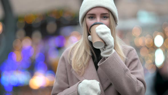 cute young girl in a hat and mittens drinks hot coffee on the background of Christmas lights