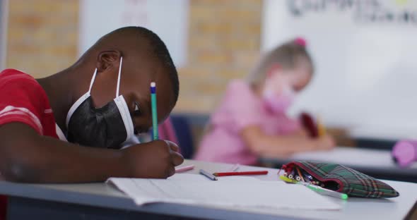 Portrait of african american schoolboy sitting in classroom, making notes wearing face mask alt