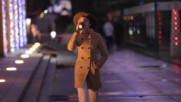 Beautiful Woman in Hat and Coat Drinks Coffee and Walks in the Night City