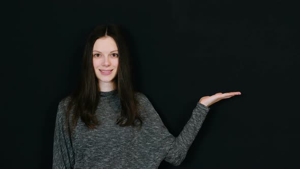 Smiling Brunette Woman Shows Hand Palm To the Right and Look on It in Black Background