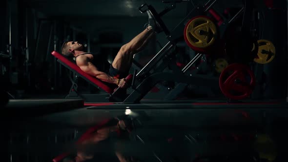 A Muscular Man Performs Exercises on a Sports Training Apparatus for Leg Muscles in a Dark Gym alt