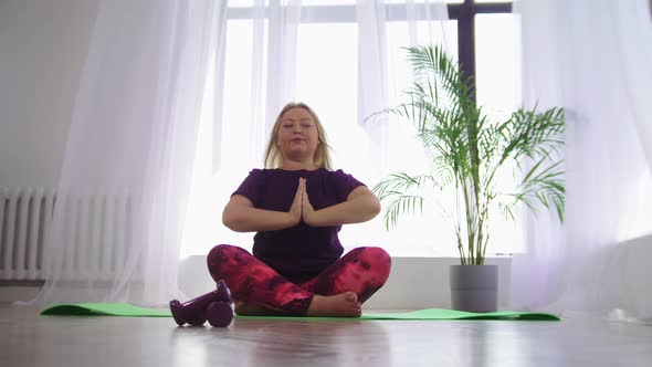 Yoga Indoors  Blonde Overweight Smiling Woman Meditating on Yoga Mat in White Modern Studio alt