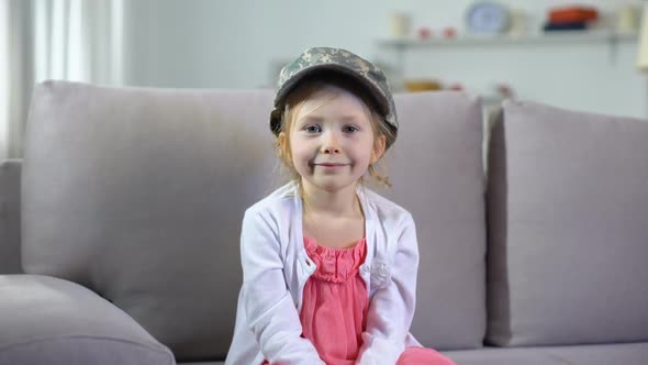 Funny Smiling Preschooler Girl in Military Cap Sitting on Sofa, Little Patriot alt