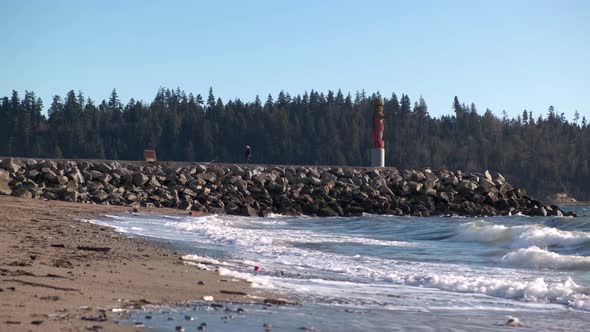 Young woman walking away from a tall statue on a rocky pier on the beach with waves hitting the shor alt