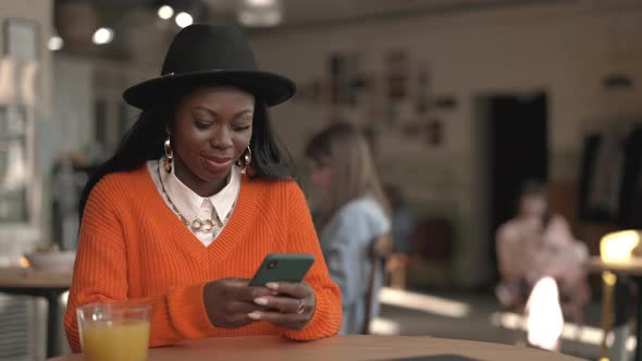 Smiling African American Woman in Trendy Black Hat Reading Messages on Smartphone While Sitting at alt