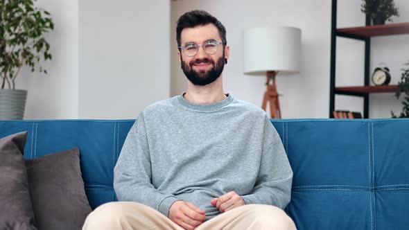 Happy Hispanic Bearded Man in Eyeglasses Relaxing on Comfortable Blue Couch Spending Time at Home alt