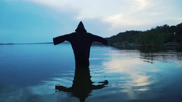 Ghost woman in the water outdoor. Scary figure in black mantle. alt