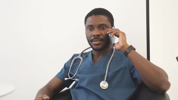 Portrait of Young AfricanAmerican Doctor Talking on Smartphone Sitting in Lobby of Modern Medical