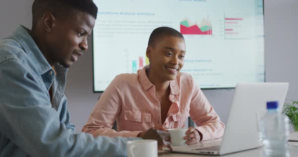 Smiling african american male and female colleague in discussion at meeting using laptop and screen alt