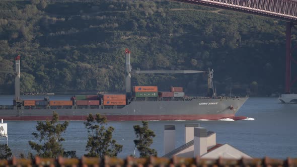 A Cargo Boat Passing Under The Abril Suspension Bridge In Portugal