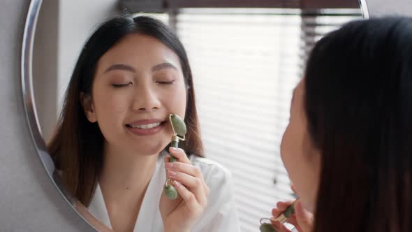 Beautiful Young Asian Woman Looking At Mirror Using Greenstone Jade Facial Roller alt