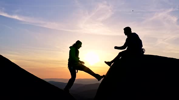 Silhouettes of a Man Helping a Woman Climbing Up at Sunset