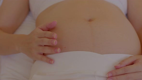 Happy Pregnant Woman sitting on bed holding and stroking her big belly at home alt