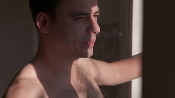 Close-up young muscular man smoking a cigarette and looks out the window alt