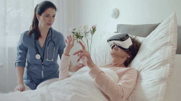 Female Doctor Checking on Elderly Patient Lying in Hospital Bed Doing Therapy Via VR Technology