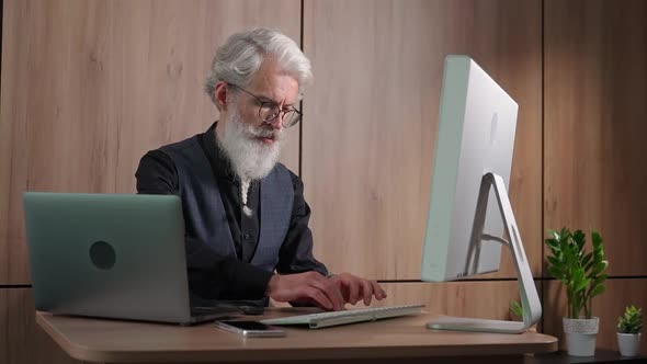 Adult Grayhaired Man Designer Works in the Office at the Computer Typing and Surfing the Internet an alt