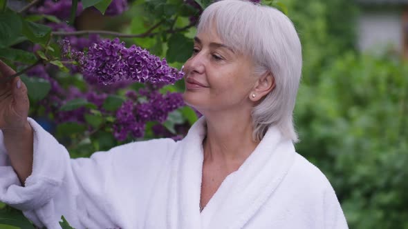 Cheerful Middle Aged Woman Smelling Purple Blossom on Tree in Garden Smiling alt