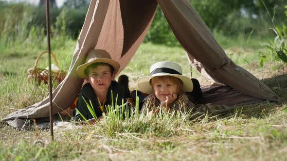 Portrait of Cute Little Children in Hat Lie on Green Lawn in a Wigwam While Resting in Countryside alt