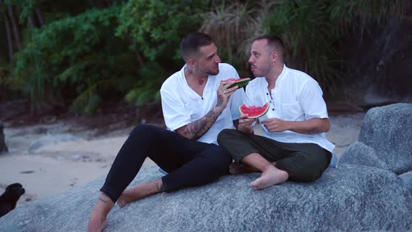 Happy Caucasian Gay Male Couple Eating Watermelon Sitting on a Rock in Tropical Island at Dusk alt