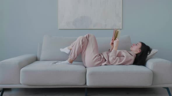 Young caucasian woman with glasses lying on sofa in cozy living room and reading book.