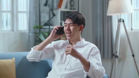 Smiling Asian Man Answering The Call On Smartphone While Lying On Sofa In The Living Room alt