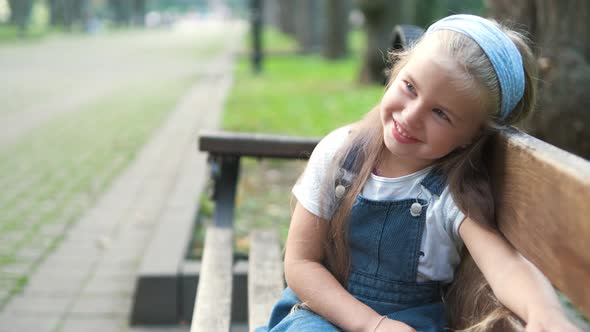 Little Happy Child Girl Sitting on a Bench Smiling Happily in Summer Park alt