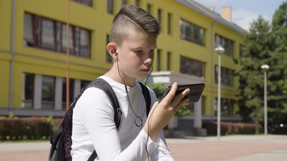 A Caucasian Teenage Boy Looks at a Smartphone with Earphones on  a School in the Background