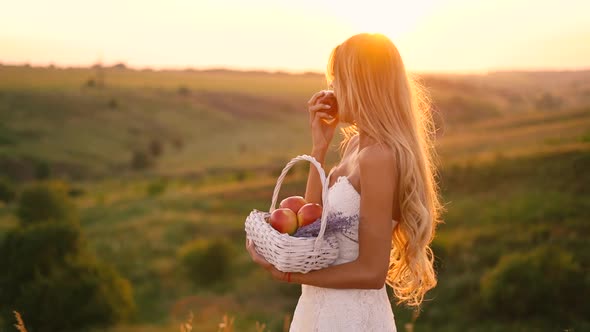Beautiful sexy blonde girl in white dress posing in a field at sunset with a basket of fruit alt