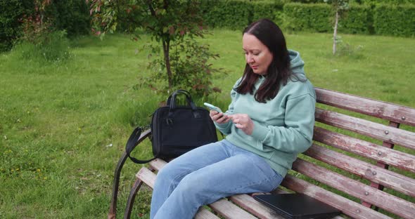 Woman Is Reading Something In Her Mobile Phone Sitting On Bench In City Park
